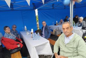 Powiększ zdjęcie: mężczyzna na wózku, za nim ludzie siedzący przy stołach pod namiotem piknikowym