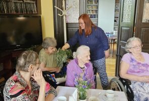 Powiększ zdjęcie: zielarka podaje kobietom przy stoliku do powąchania gałązki piołunu