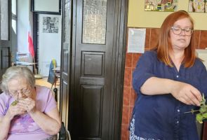 Powiększ zdjęcie: zielarka z ziołami w rękach, obok kobieta wącha zioła