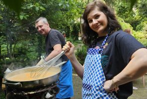 Powiększ zdjęcie: kobieta smażąca jajecznicę na dużej patelni, za nią mężczyzna przygotowujący jajka