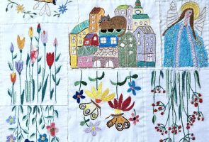 Powiększ zdjęcie: praca złożona z haftów przedstawiających kwiaty, rybę, domy i anioła
