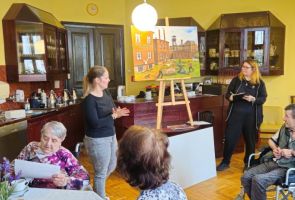 Powiększ zdjęcie: duży kolorowy obraz na sztaludze , obok dwie kobiety oraz nasi Mieszkańcy  w kawiarni dps