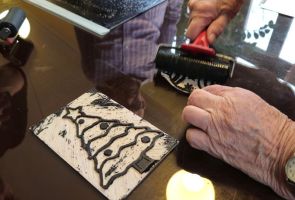 Powiększ zdjęcie: dłonie przenoszące farbę wałkiem na linoryt, obok linoryt z choinką