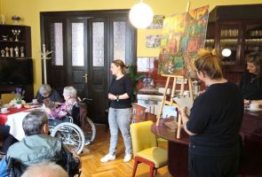 Powiększ zdjęcie: duży kolorowy obraz na sztaludze , obok dwie kobiety oraz nasi Mieszkańcy  w kawiarni dps