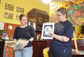 Powiększ zdjęcie: duży kolorowy obraz na sztaludze , obok dwie kobiety w kawiarni dps