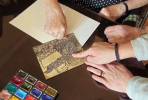 Powiększ zdjęcie: dłonie wskazujące linoryt leżący na stoliku, obok kartka papieru i farby