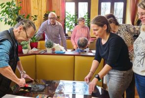 Powiększ zdjęcie: kobieta rozprowadzająca wałkiem farbę na szybie, wokół obserwują ją uczestnicy warsztatów