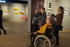 Powiększ zdjęcie: nasi Mieszkańcy słuchający oprowadzającej po wystawie kobiety